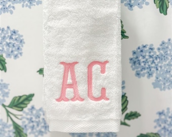 May include: White hand towel with pink embroidered "AC" initials. The towel is hanging against a backdrop of a floral pattern with blue flowers and green leaves.