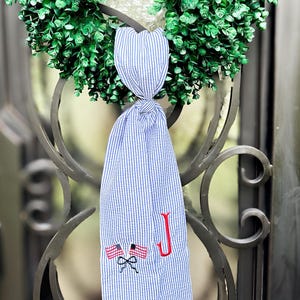 May include: A green wreath with a blue and white striped fabric hanging from it. The fabric has an embroidered red letter "J" and two American flags.