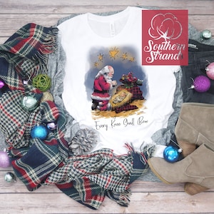 May include: White long-sleeve t-shirt with a watercolor illustration of Santa kneeling before baby Jesus in a manger. The text "Every Knee Shall Bow" is printed below the image.