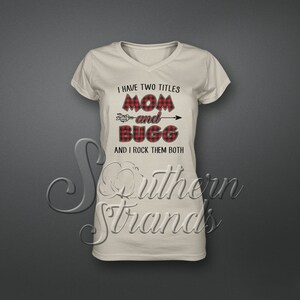 May include: A beige v-neck t-shirt with the text "I have two titles Mom and Bugg and I rock them both" in black text with a red and black plaid design behind the text.