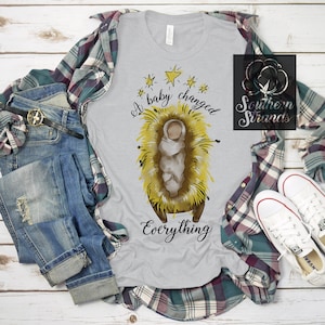 May include: A light grey t-shirt featuring a graphic of a baby in a manger, with yellow straw and stars. The text "A baby changed Everything" is written in black script. The shirt is styled with blue jeans, a plaid shirt, and white trainers.