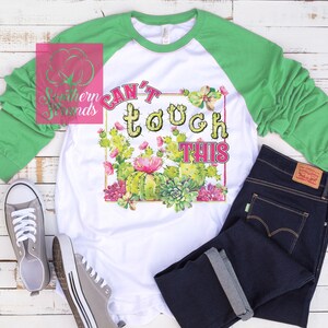 May include: White and green raglan shirt with the text "Can't Touch This" in a playful font, surrounded by a cactus and floral design. The shirt is paired with dark wash jeans and gray sneakers, creating a casual, everyday look.