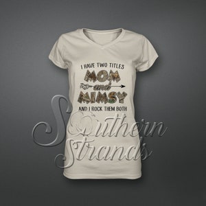 Op de afbeelding: Een beige V-hals T-shirt met een camouflageprint. Het shirt heeft de tekst "I have two titles Mom and Mimsy and I rock them both" erop gedrukt.