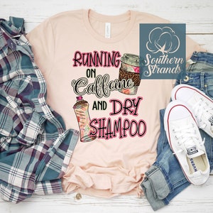 May include: A light pink t-shirt with the text "Running on Caffeine and Dry Shampoo" in a playful font. The shirt features graphic illustrations of a coffee cup and a dry shampoo bottle. A plaid shirt, jeans, and white sneakers complete the casual look.
