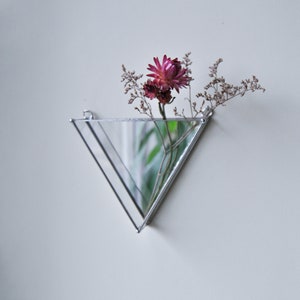 May include: A triangular wall-mounted mirror with a silver frame holds a single pink flower and dried foliage.