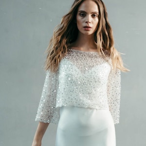 May include: A white, beaded, off-the-shoulder top with long sleeves and a flowing skirt. The model is wearing gold sandals.