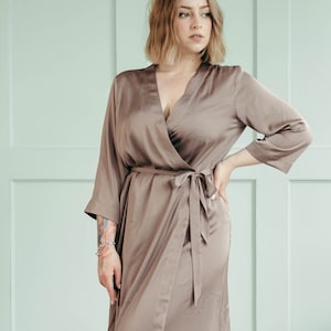 May include: A woman wearing a long, brown satin robe with a wrap style and a V-neck. The robe has a sash tied at the waist.