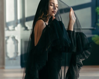 May include: A woman wearing a black tulle dress and veil. The veil is covering her face and the dress has ruffles.
