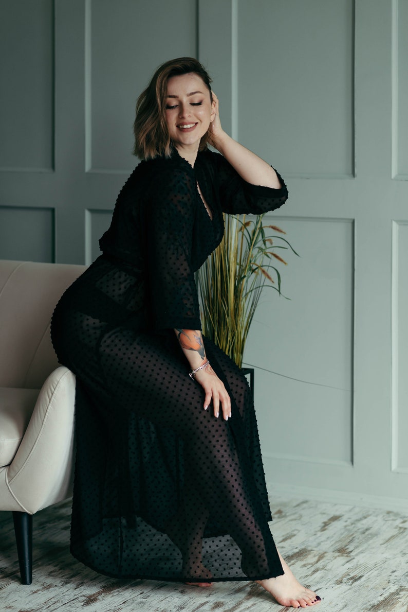 May include: A woman in a black sheer polka dot robe sits on a white couch. She is smiling and looking down.