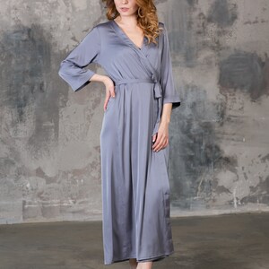 May include: A long, grey satin robe with a wrap closure and a tie belt. The robe is worn by a person standing in front of a grey concrete wall.