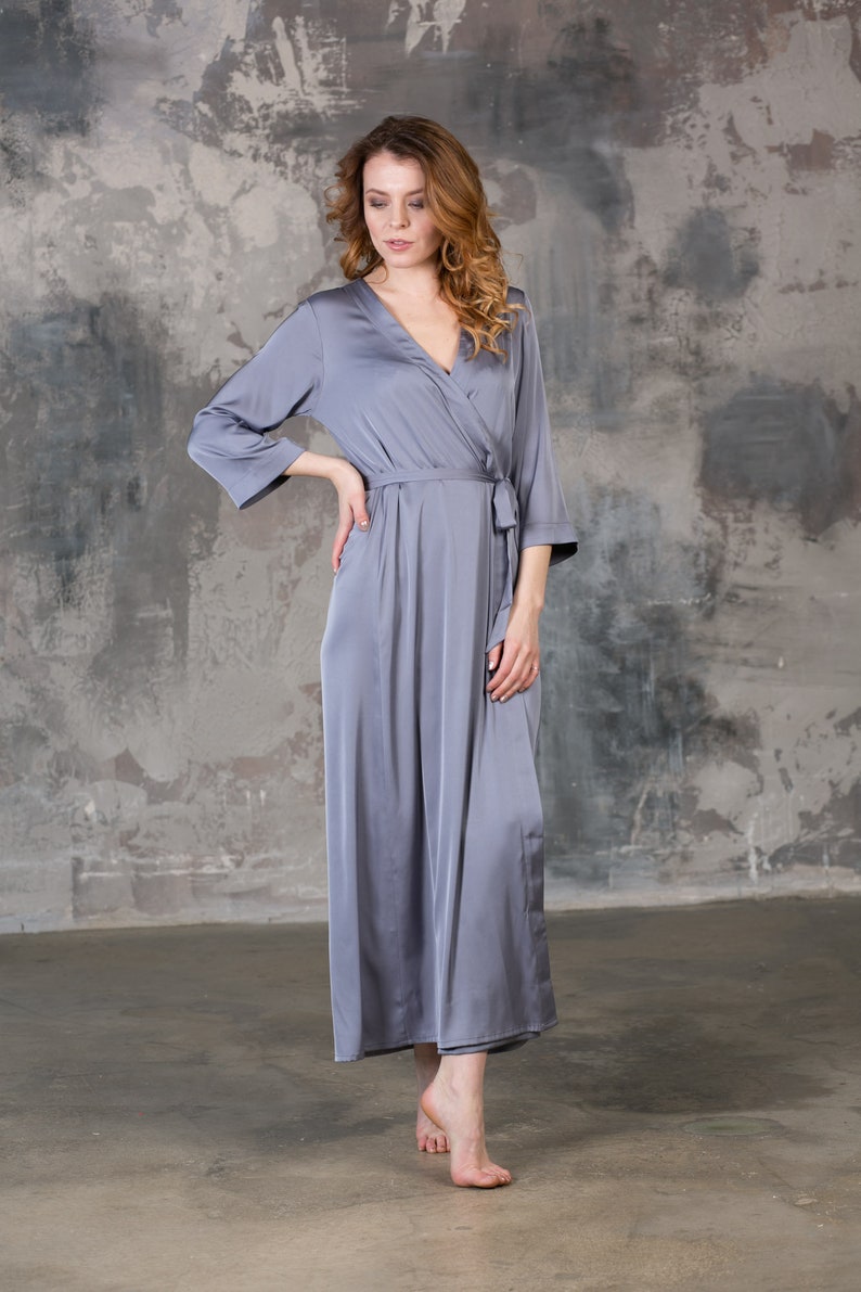 May include: A long, grey satin robe with a wrap closure and a tie belt. The robe is worn by a person standing in front of a grey concrete wall.