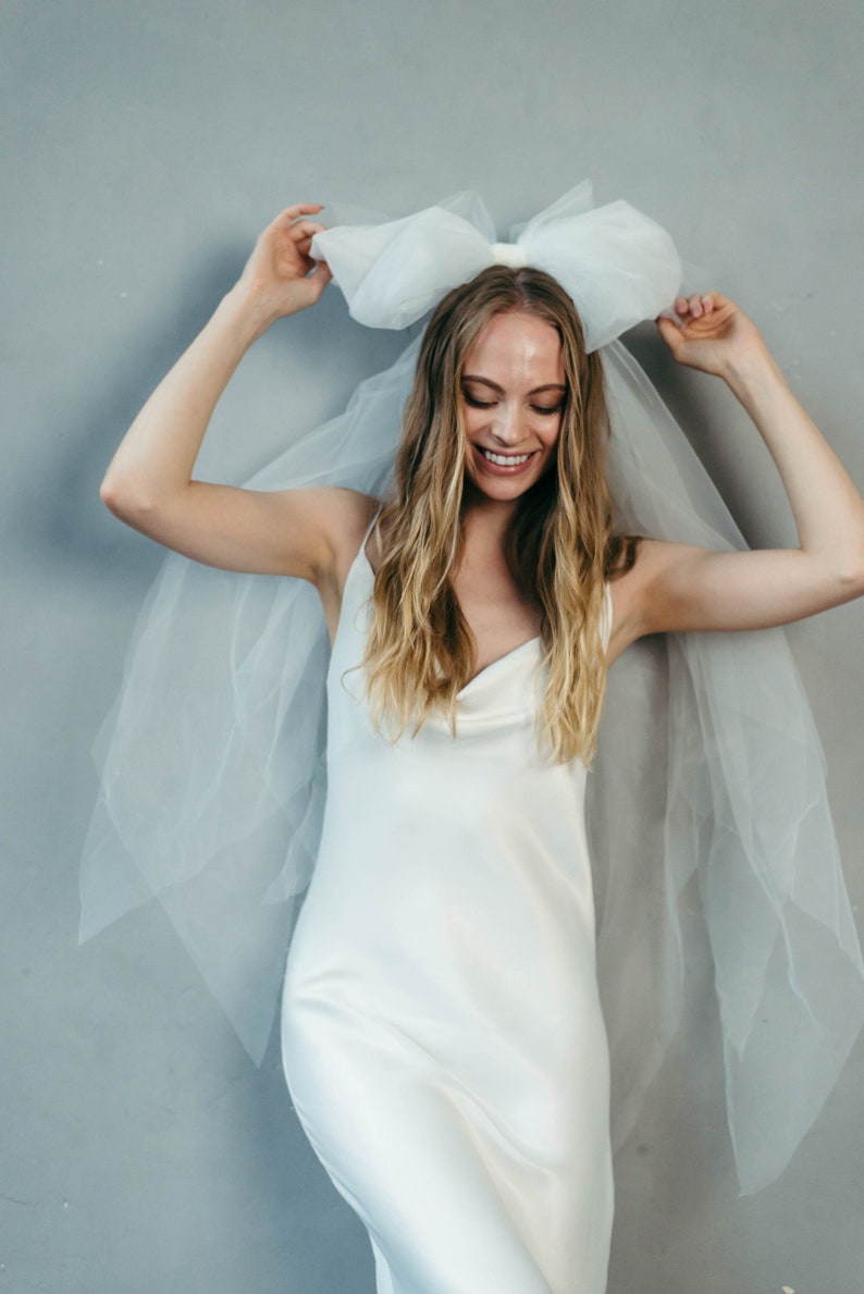 May include: A woman wearing a white satin dress with a large white tulle bow on her head. The bow is attached to a veil that drapes down her back.