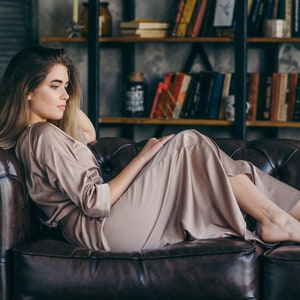 May include: A woman wearing a light brown satin robe is lying on a brown leather sofa. The sofa has a tufted design and is in a room with a bookcase in the background.
