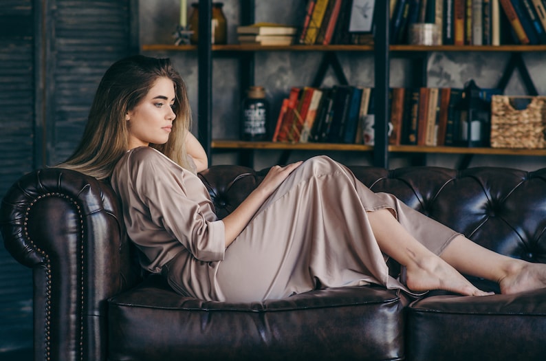 May include: A woman wearing a light brown satin robe is lying on a brown leather sofa. The sofa has a tufted design and is in a room with a bookcase in the background.