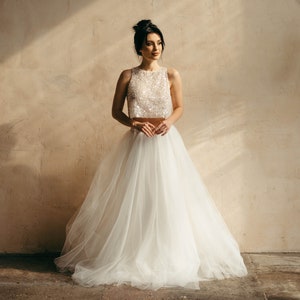 May include: A woman wearing a white, beaded crop top and a flowing tulle skirt. The skirt is very full and has a lot of movement. The woman is standing in front of a beige wall.