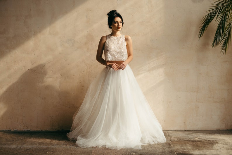 May include: A woman wearing a white, beaded crop top and a flowing tulle skirt. The skirt is very full and has a lot of movement. The woman is standing in front of a beige wall.