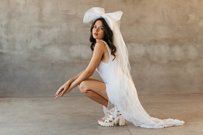 May include: A woman wearing a white dress and a large white bow on her head. She is wearing white heels and has her hair down. She is sitting on the floor with her legs crossed.