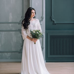 Op de afbeelding: Een vloerlange witte jurk met een kanten lijfje met driekwartmouwen en een effen, zwierige rok. Het kledingstuk wordt gecombineerd met een boeket witte rozen en groen blad, dat op taillehoogte in een staande positie wordt vastgehouden.