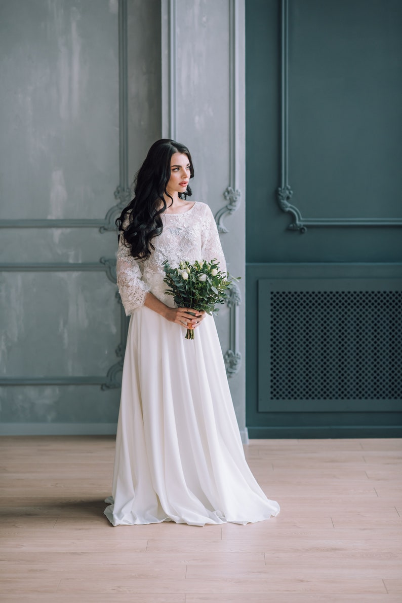 Op de afbeelding: Een vloerlange witte jurk met een kanten lijfje met driekwartmouwen en een effen, zwierige rok. Het kledingstuk wordt gecombineerd met een boeket witte rozen en groen blad, dat op taillehoogte in een staande positie wordt vastgehouden.