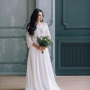 Op de afbeelding: Een vloerlange witte jurk met een kanten lijfje met lange mouwen, een ronde halslijn en een zwierige, effen witte rok. Het kledingstuk wordt gecombineerd met een boeket witte rozen en groen blad dat in het midden wordt vastgehouden.