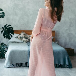 Op de afbeelding: Een doorschijnende, lichtroze robe met een stippenpatroon. De robe heeft lange mouwen en een bijpassende riem. Het model staat in een slaapkamer met een bed en decoratieve planten op de achtergrond.
