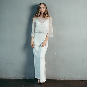 May include: A white, long-sleeved, beaded top with a matching white satin skirt. The top has a scoop neckline and a loose, flowing fit. The skirt is fitted at the waist and flows to the floor.