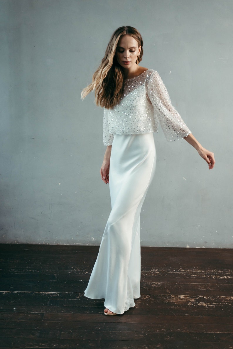 May include: A white, sequined, semi-sheer top with long sleeves and a scoop neck, paired with a white, flowing, satin skirt.