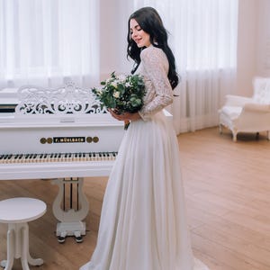 Op de afbeelding: Een vloerlange witte jurk met lange kanten mouwen en een effen rok, gecombineerd met een boeket witte bloemen en groen blad. Het kledingstuk heeft een hoge halslijn en een aansluitend lijfje dat overgaat in een zwierige, lange rok.