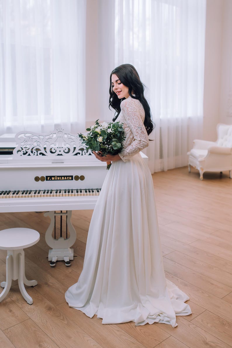 Op de afbeelding: Een vloerlange witte jurk met lange kanten mouwen en een effen rok, gecombineerd met een boeket witte bloemen en groen blad. Het kledingstuk heeft een hoge halslijn en een aansluitend lijfje dat overgaat in een zwierige, lange rok.