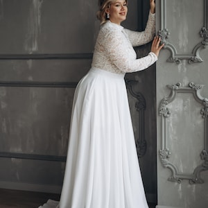 Op de afbeelding: Een vloerlange witte jurk met een kanten lijfje met lange mouwen, een hoge halslijn en een effen, zwierige rok. Het kledingstuk heeft een getailleerde pasvorm en een rok van gladde, ondoorzichtige stof, wat contrasteert met het gedetailleerde bloemenkantpatroon op het bovenstuk.
