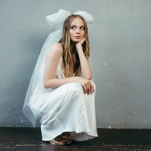 May include: A woman in a white satin dress with a large white bow on her head. She is sitting on the floor with her legs crossed and her chin resting on her hand.