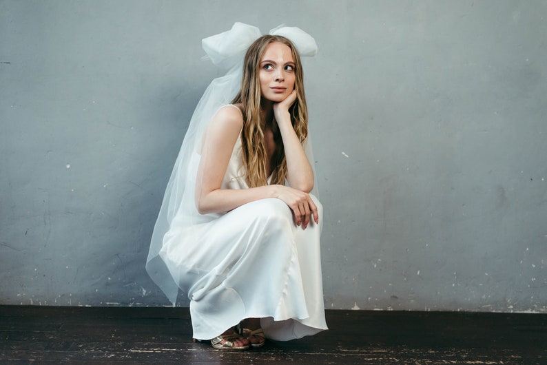 May include: A woman in a white satin dress with a large white bow on her head. She is sitting on the floor with her legs crossed and her chin resting on her hand.