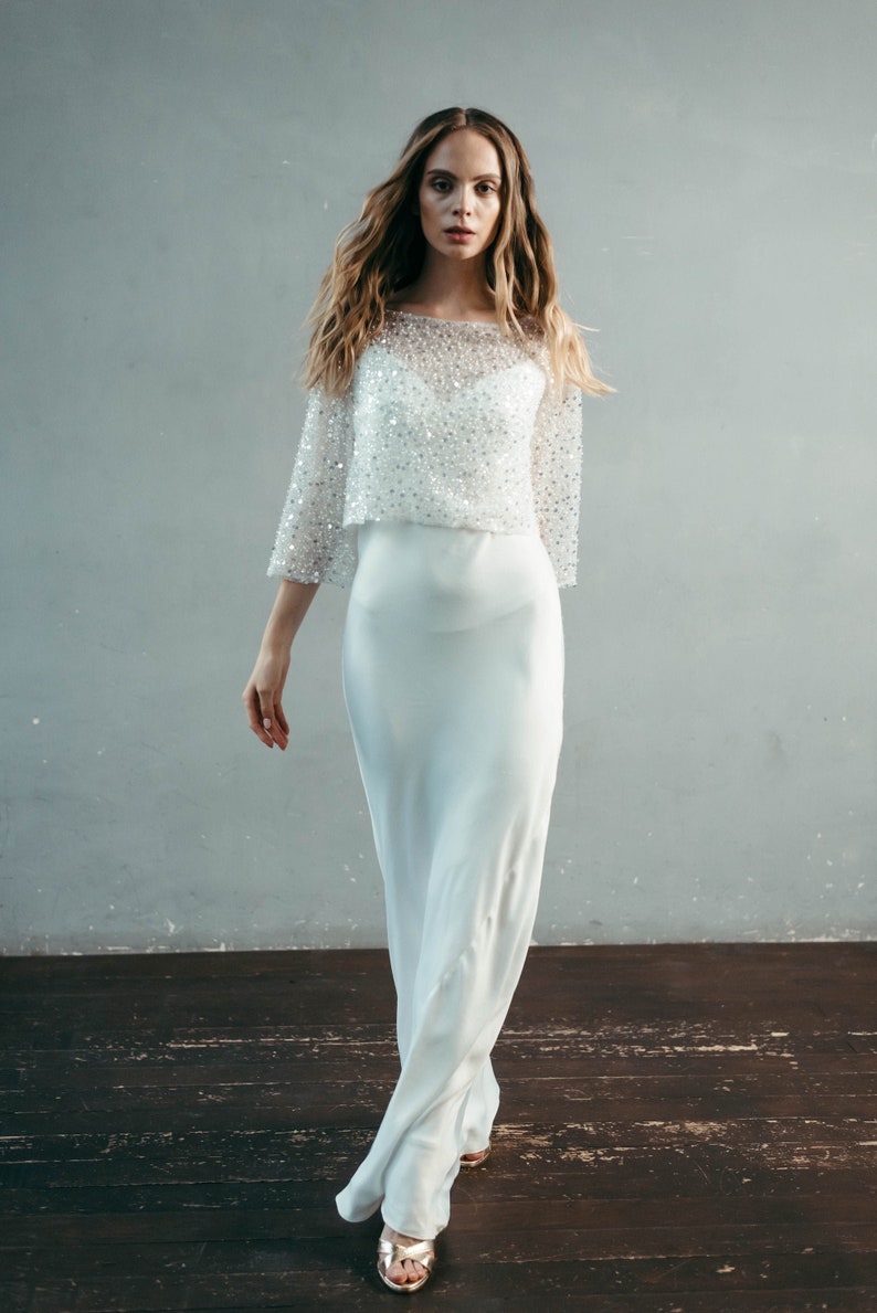 May include: A white, beaded, off-the-shoulder top with long sleeves and a flowing skirt. The model is wearing gold sandals.