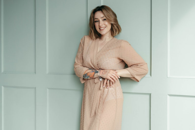 May include: A woman with short brown hair is wearing a light brown, sheer, polka dot dress with a V-neckline. She is standing in front of a light green wall with white trim.