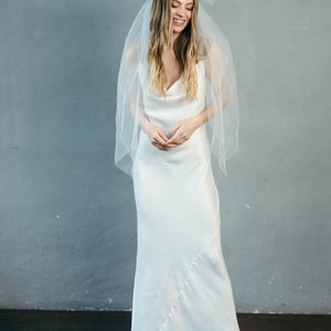 May include: A woman wearing a white satin wedding dress with a cowl neck and a white tulle veil with a large bow. She is smiling and looking down.