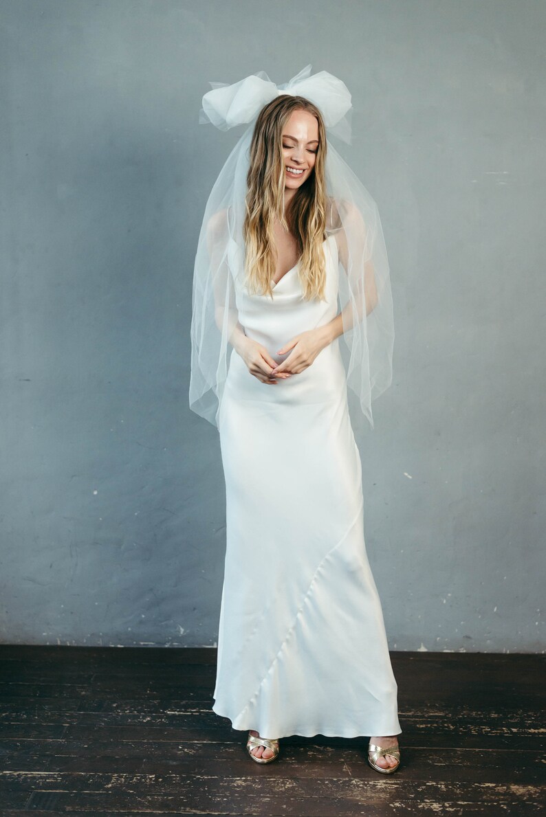 May include: A woman wearing a white satin wedding dress with a cowl neck and a white tulle veil with a large bow. She is smiling and looking down.