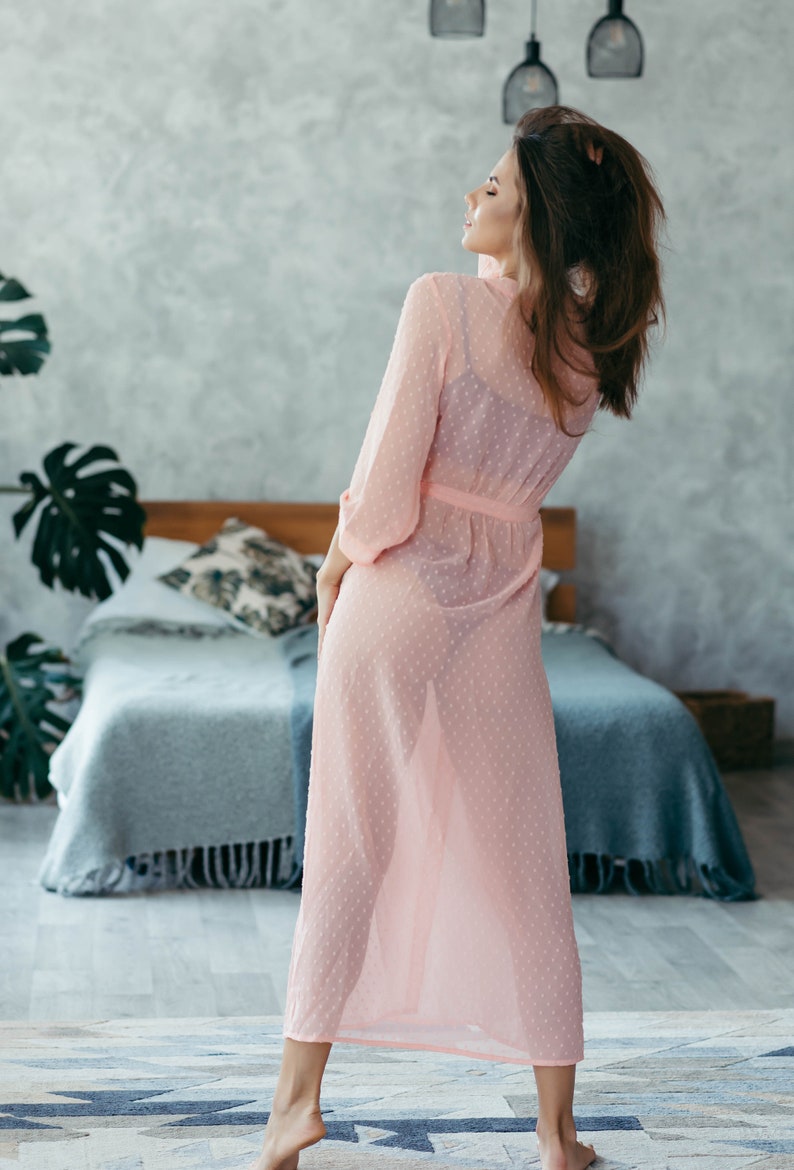May include: A woman in a pink sheer robe with white polka dots stands in a bedroom with a bed behind her. The robe is tied at the waist and has long sleeves.