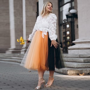 Könnte beinhalten: Eine Frau trägt eine weiße Spitzenbluse und einen mehrfarbigen Tüllrock. Der Rock ist in orangefarbene und schwarze Abschnitte unterteilt. Sie trägt goldene High Heels und hält gelbe Blumen. Der Hintergrund zeigt ein Gebäude mit Säulen.