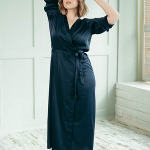 A bridesmaid wearing a long, dark navy blue satin robe with a tie belt. She is standing in a room with white walls and wooden floors.