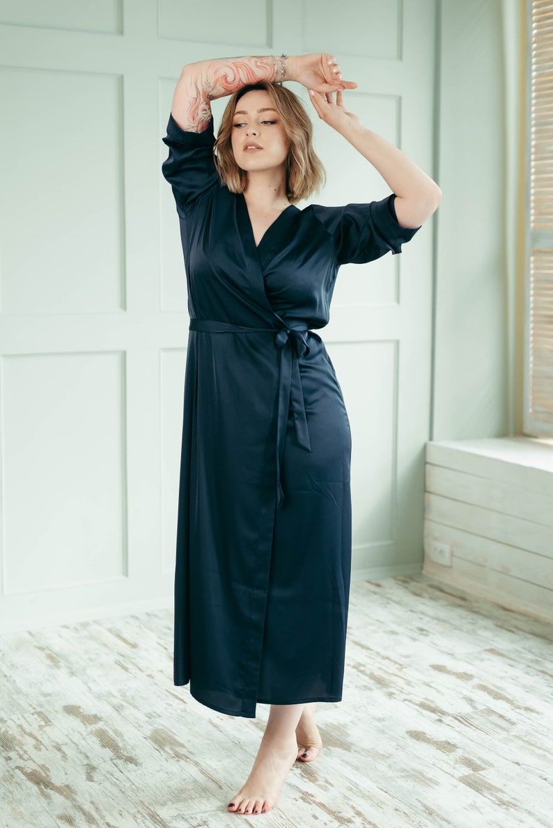 A bridesmaid wearing a long, dark navy blue satin robe with a tie belt. She is standing in a room with white walls and wooden floors.