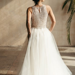May include: A woman wearing a white tulle skirt and a sequined top. The skirt is flowing and the top is sleeveless. The woman is standing in a room with a concrete floor and a wall in the background.