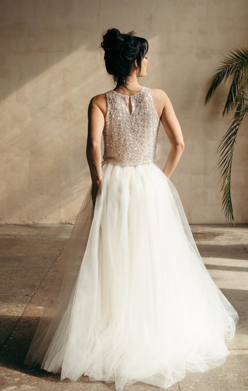 May include: A woman wearing a white tulle skirt and a sequined top. The skirt is flowing and the top is sleeveless. The woman is standing in a room with a concrete floor and a wall in the background.