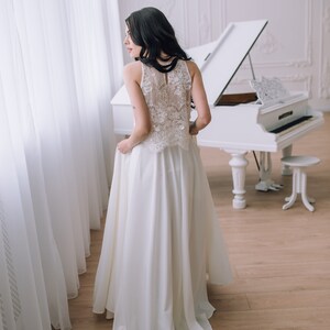 Op de afbeelding: Een vloerlange witte jurk met een mouwloos lijfje met gedetailleerde bloemenkant en een hoge halslijn. De rok is gemaakt van een effen, gladde stof die tot op de grond valt en een strakke en elegante silhouet cre&euml;ert.