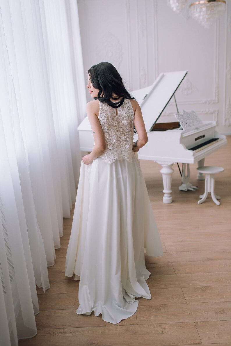 Op de afbeelding: Een vloerlange witte jurk met een mouwloos lijfje met gedetailleerde bloemenkant en een hoge halslijn. De rok is gemaakt van een effen, gladde stof die tot op de grond valt en een strakke en elegante silhouet cre&euml;ert.