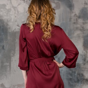 May include: A long, burgundy satin robe with a tie belt. The robe is being worn by a person with long, curly hair.