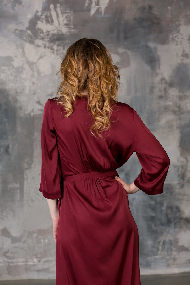 May include: A long, burgundy satin robe with a tie belt. The robe is being worn by a person with long, curly hair.