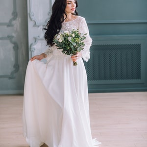 Op de afbeelding: Een vloerlange witte jurk met een kanten lijfje met driekwartmouwen en een effen, vloeiende rok. Het kledingstuk wordt gecombineerd met een boeket witte rozen en groene eucalyptusbladeren, vastgehouden voor het middel.