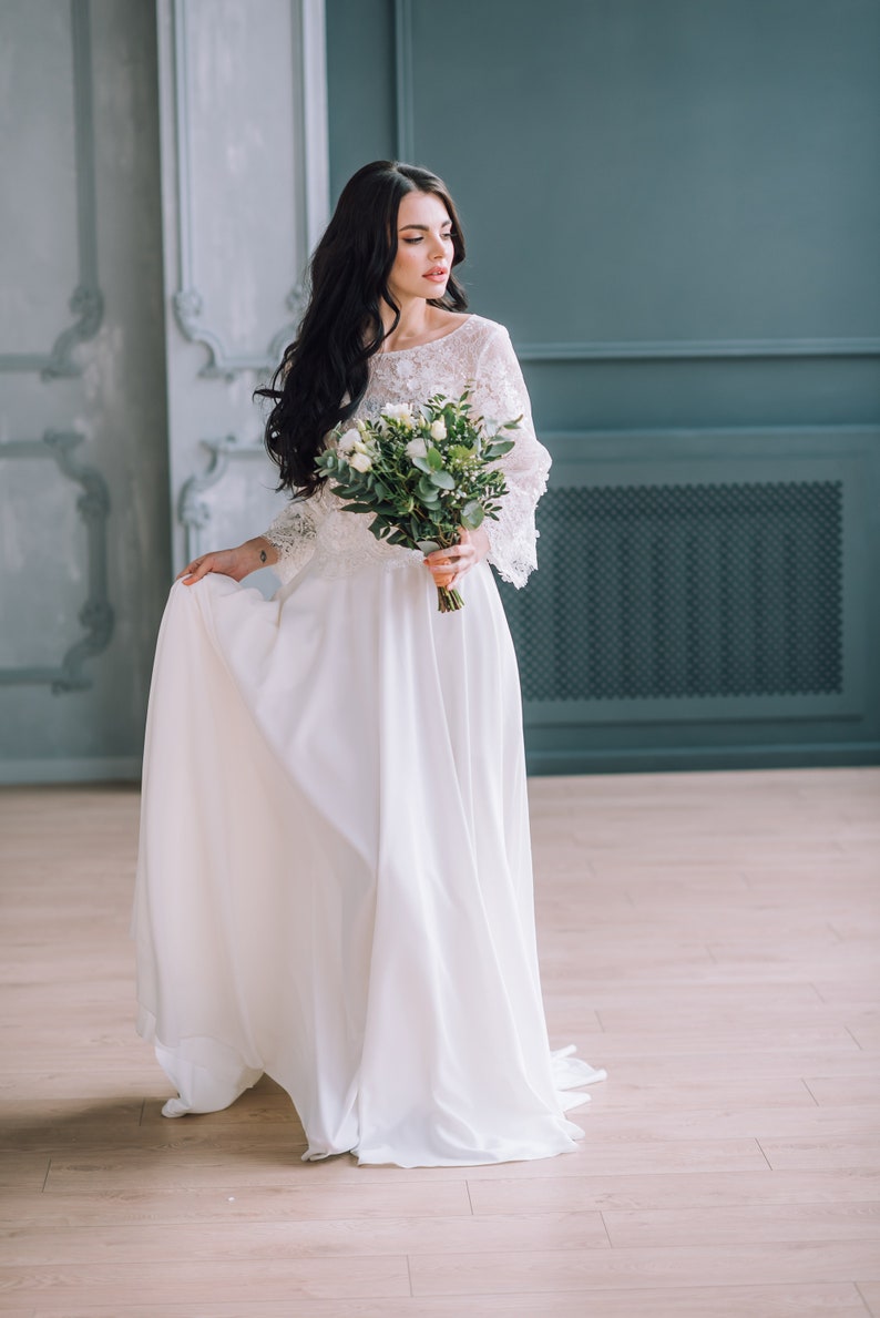 Op de afbeelding: Een vloerlange witte jurk met een kanten lijfje met driekwartmouwen en een effen, vloeiende rok. Het kledingstuk wordt gecombineerd met een boeket witte rozen en groene eucalyptusbladeren, vastgehouden voor het middel.