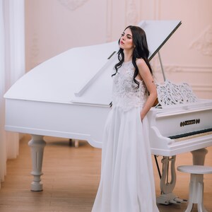 Op de afbeelding: Een vloerlange witte jurk met een mouwloos kanten lijfje met een hoge halslijn en een effen witte rok met zijzakken. Het kledingstuk is geplaatst voor een witte vleugel met een bijpassende witte pianobank op een houten vloer.