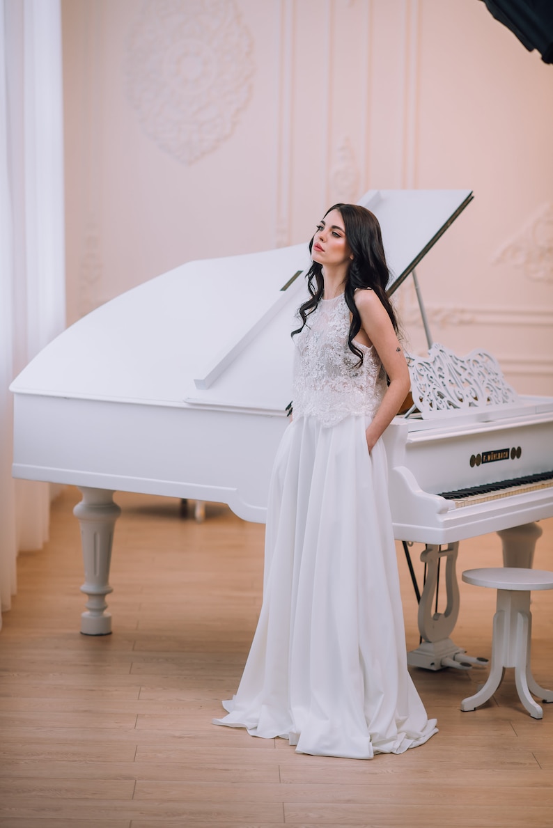 Op de afbeelding: Een vloerlange witte jurk met een mouwloos kanten lijfje met een hoge halslijn en een effen witte rok met zijzakken. Het kledingstuk is geplaatst voor een witte vleugel met een bijpassende witte pianobank op een houten vloer.